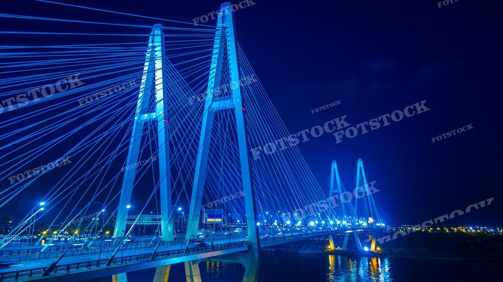 На фотографии ночной вид на ярко освещённый вантовый мост, подсвеченный синими огнями, которые отражаются на водной поверхности. Эффектная подсветка и геометрические линии вантов создают впечатление динамики и современности. Изображение может быть использовано в рекламных кампаниях, урбанистических исследованиях, для декоративных целей или как образец современной инженерии и ночной архитектуры в учебных материалах.